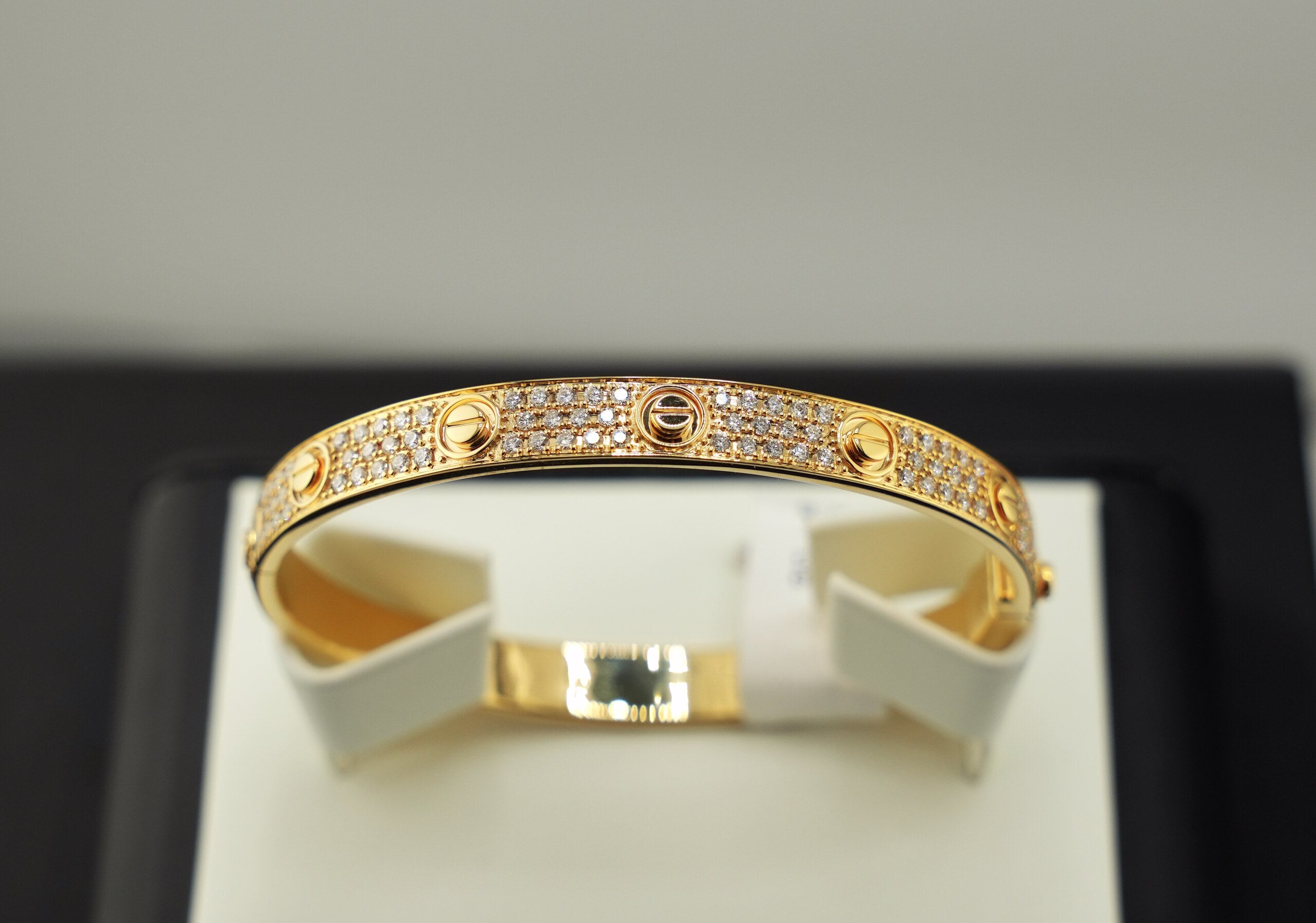 A gold bangle bracelet with pavé-set clear stones and signature screw details, displayed on a white jewelry stand against a black and light gray background.