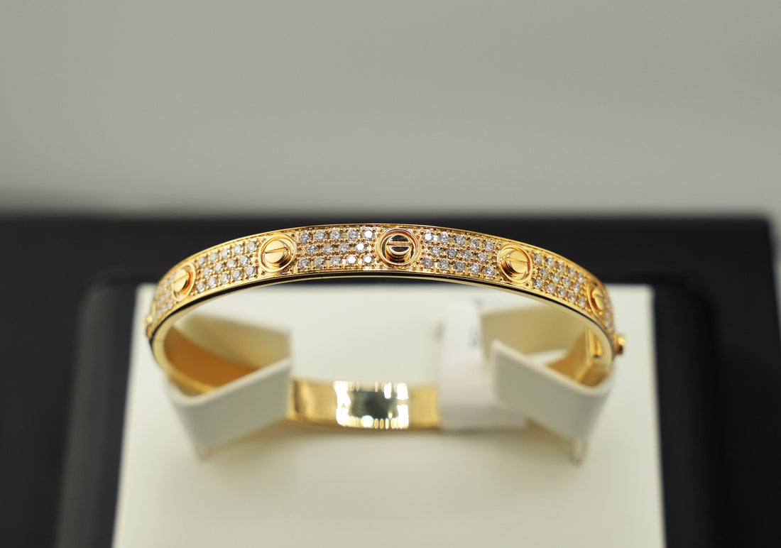 A gold bangle bracelet with pavé-set clear stones and signature screw details, displayed on a white jewelry stand against a black and light gray background.